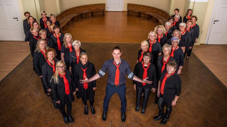 Gruppenbild der Sängerinnen mit ihrem Chorleiter in einem Foyer. Gruppenbild der Sängerinnen mit ihrem Chorleiter in einem Foyer.