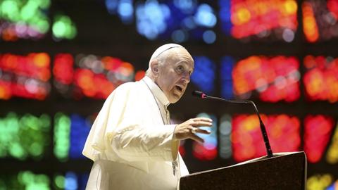 Papst Franziskus an einem Rednerpult, im Hintergrund bunte Lichter.
