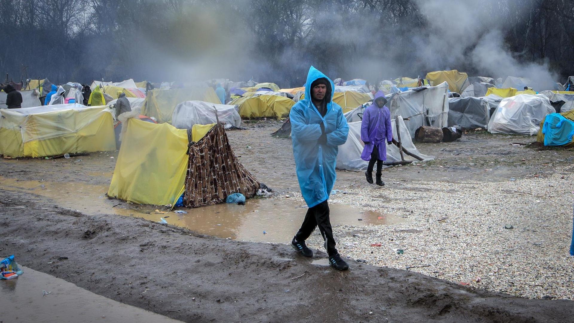 Zwei Männer im Regenponcho gehen durch ein notdürftiges Zeltlager von Flüchtlingen.