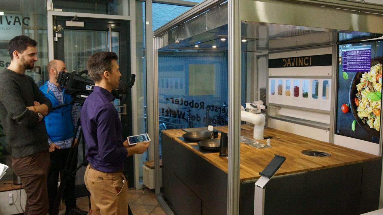 Ein Mensch mit Tablet udn ein Kamera-Team stehen vor dem Kochroboter DaVinci. Ein Mensch mit Tablet udn ein Kamera-Team stehen vor dem Kochroboter DaVinci.
