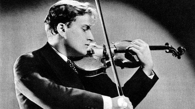 Eine alte schwarz-weiß Fotografie zeigt einen jungen Mann in schwarzem Anzug und Krawatte, der auf einer Geige einen hohen Ton greift und auf seine Finger schaut.