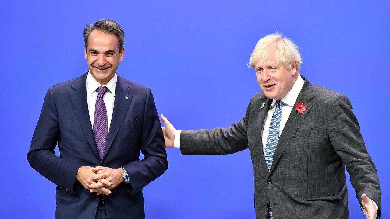 Ministerpräsident Kyriakos Mitsotakis steht im dunklen Anzug vor leuchtend blauem Hintergrund und wird vom britischen Premierminister Boris Johnson begrüßt.