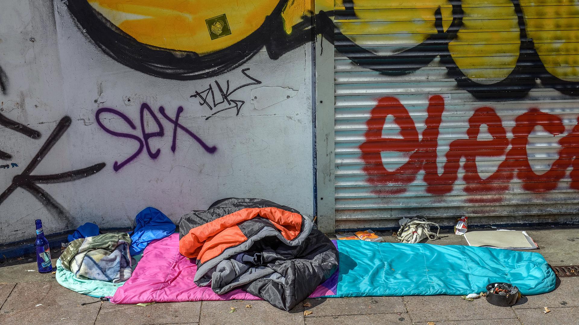 Ein Schlafsack und eine Jacke liegen vor einer Wand und einem Rolladen mit Graffitis. Ein Schlafsack und eine Jacke liegen vor einer Wand und einem Rolladen mit Graffitis.