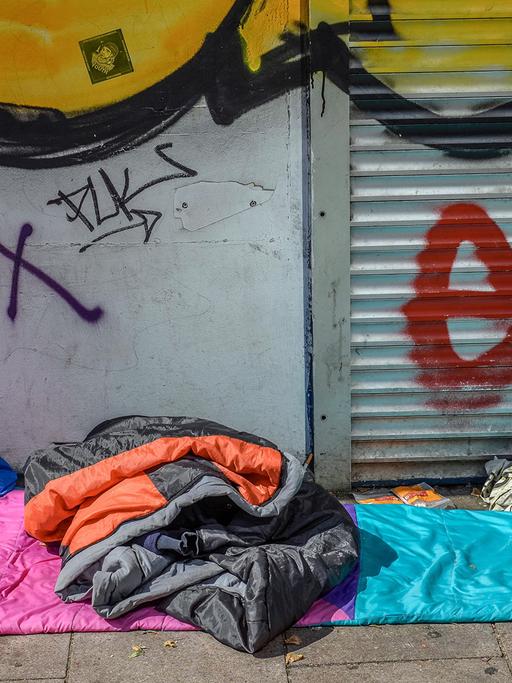 Ein Schlafsack und eine Jacke liegen vor einer Wand und einem Rolladen mit Graffitis.