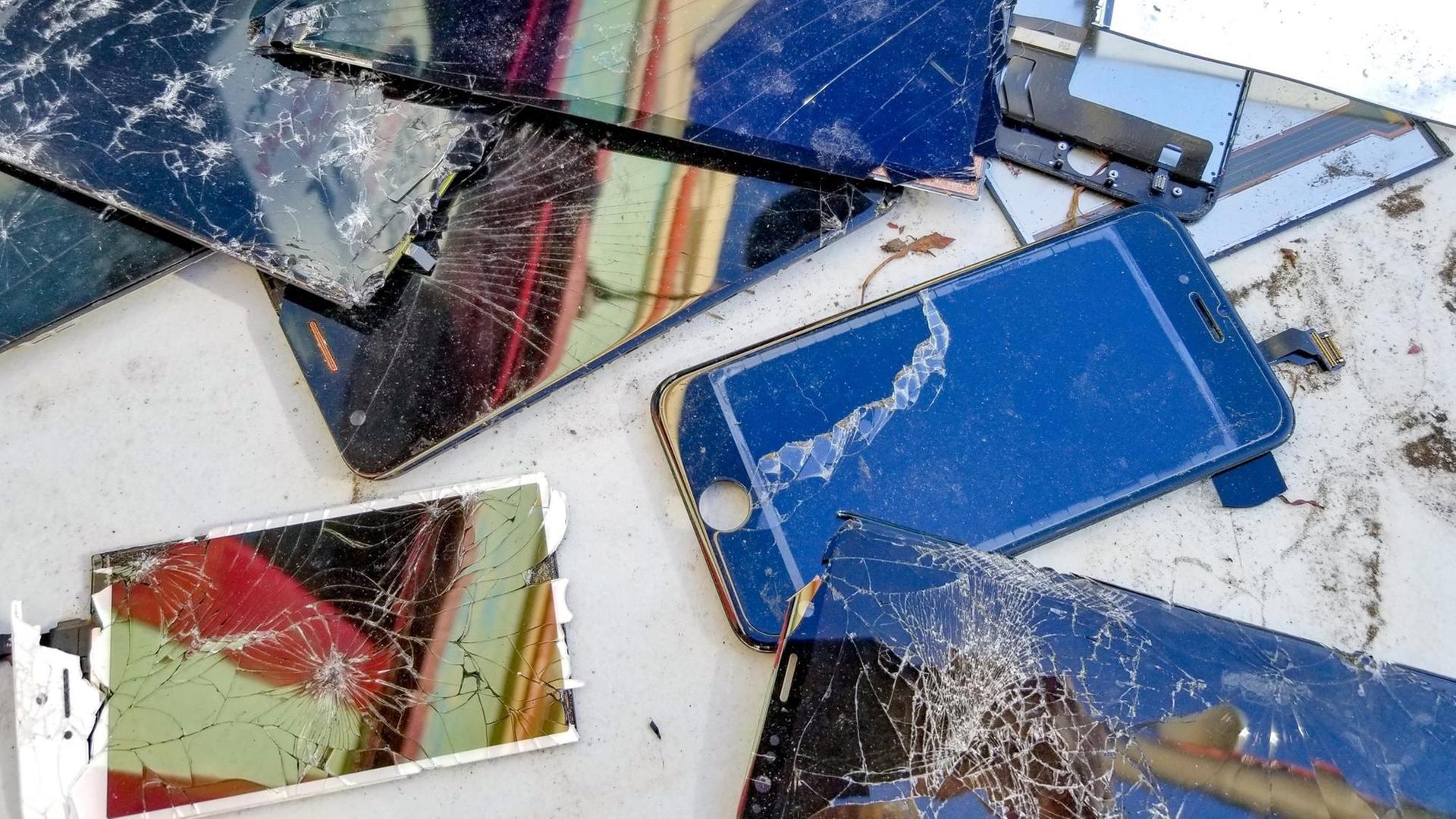 Verschiedene Smartphones mit zersprungenen Bildschirmen liegen auf einer weißen Fläche.