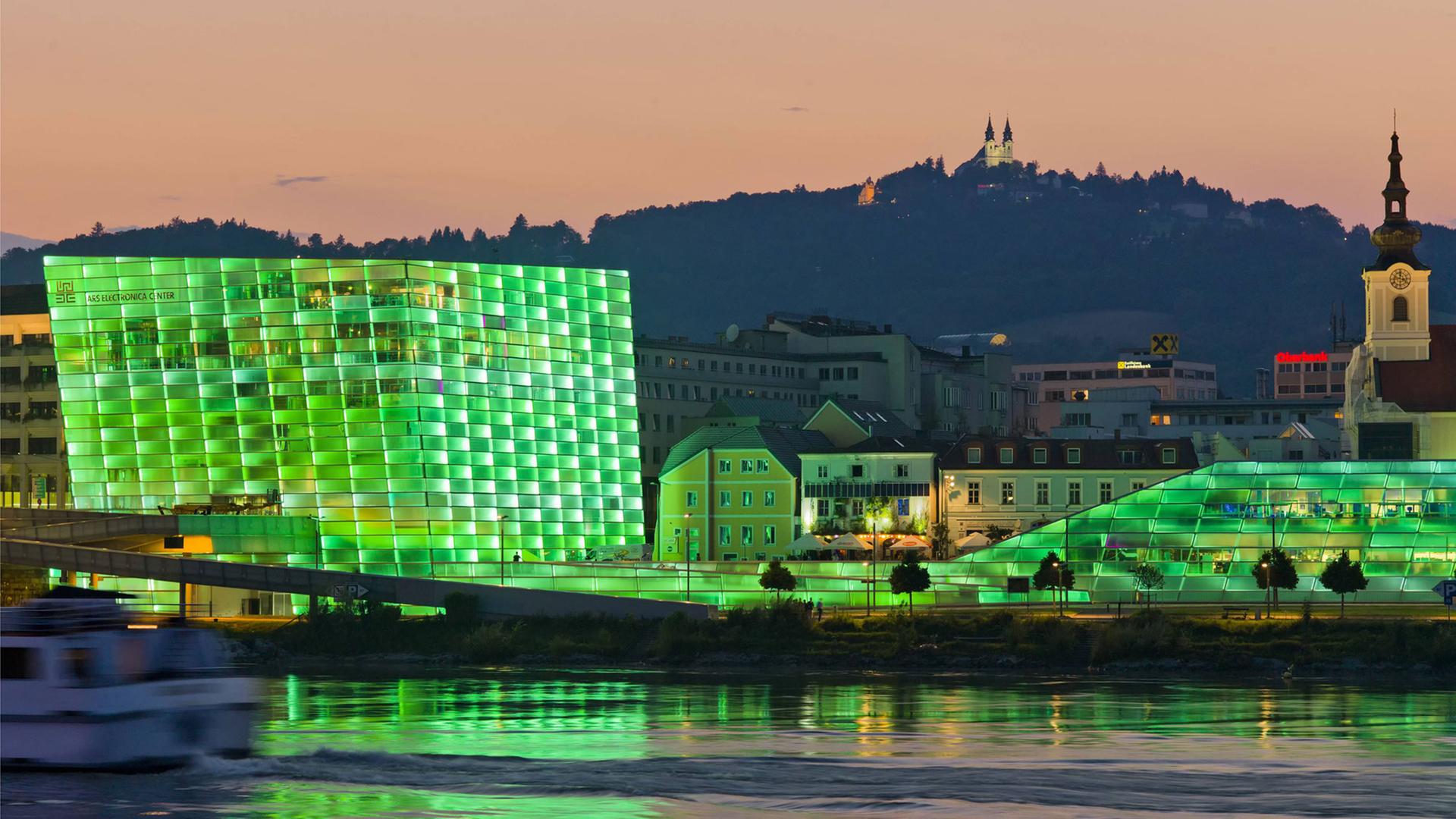 Blick auf das Ars Electronica Center in Linz mit seiner in grünes Licht getauchten Fassade. Das Gebäude spiegelt sich im Wasser, im Hintergrund ist ein Berg zu sehen. Blick auf das Ars Electronica Center in Linz mit seiner in grünes Licht getauchten Fassade. Das Gebäude spiegelt sich im Wasser, im Hintergrund ist ein Berg zu sehen.