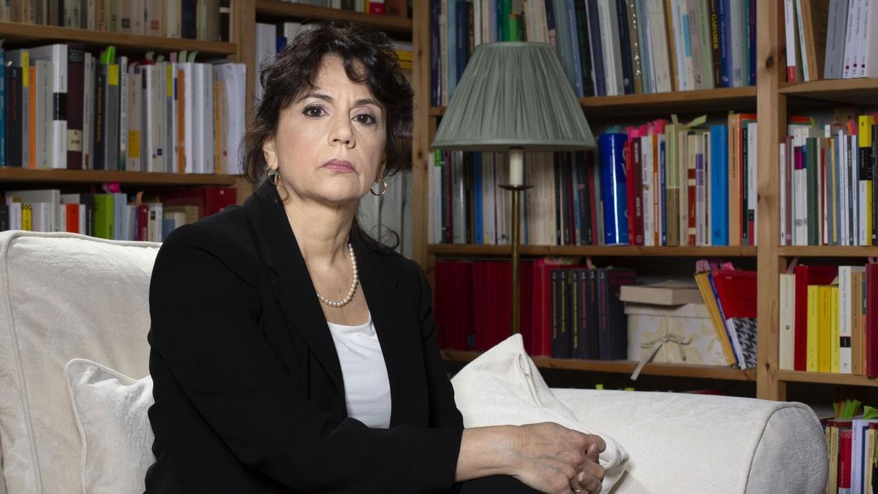 Donatella Di Cesare in einem schwarzen Sakko und weißer Bluse sitzt auf dem Sofa vor Bücherregalen, die bis zur Zimmerdecke reichen.