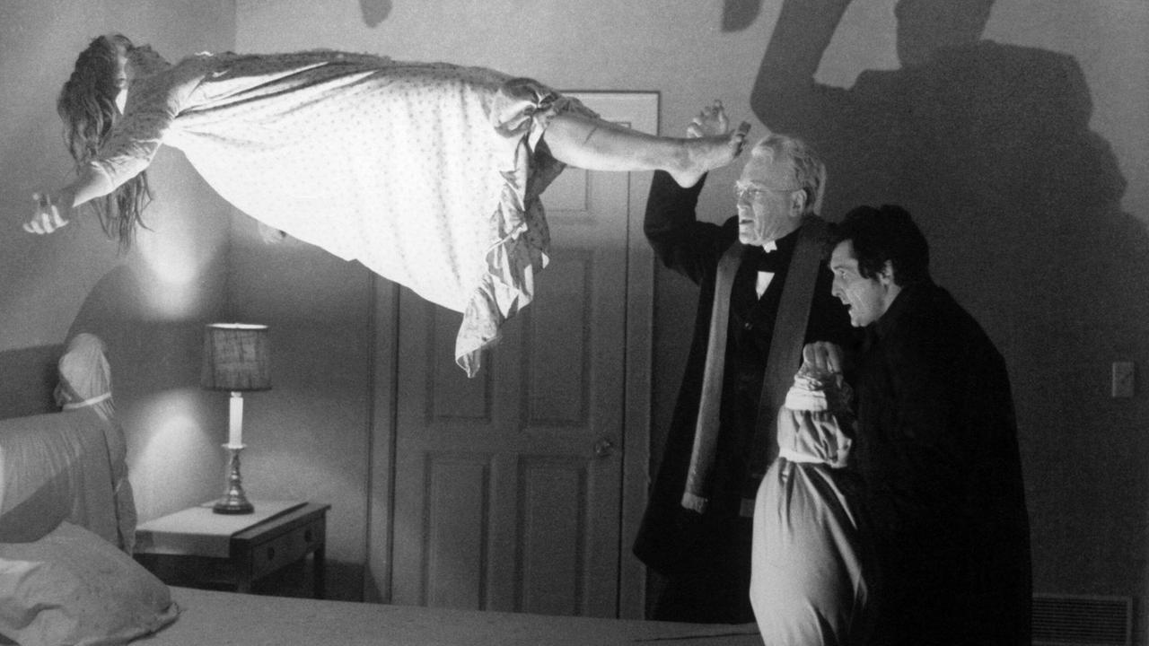 Filmstill aus "Der Exorzist" von 1973. Der Priester steht vor dem Bett des besessenen Mädchens, das in der Luft schwebt.