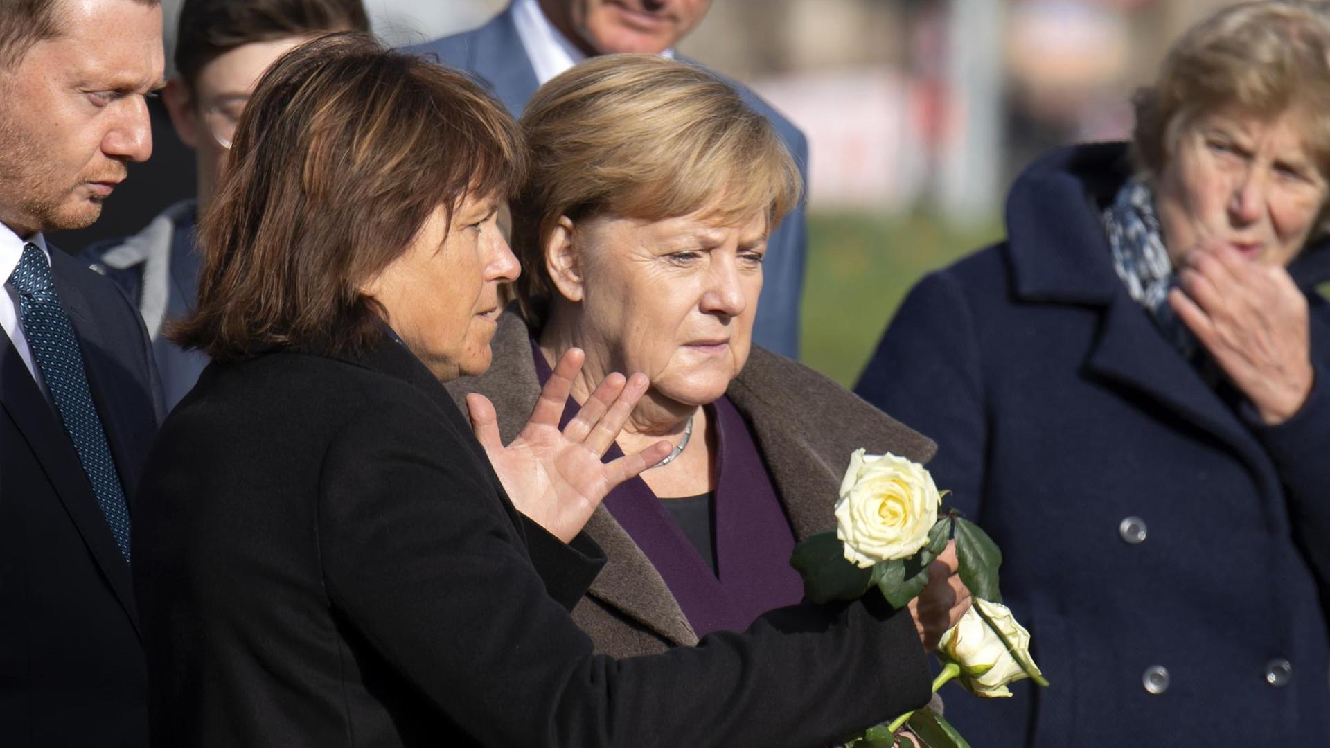 Die drei stehen nebeneinander, Merkel und Findeiß halten weiße Rosen in den Händen. Findeiß sagte etwas zu Merkel und gestikuliert mit der anderen Hand. Dahinter weitere Personen. Die drei stehen nebeneinander, Merkel und Findeiß halten weiße Rosen in den Händen. Findeiß sagte etwas zu Merkel und gestikuliert mit der anderen Hand. Dahinter weitere Personen.
