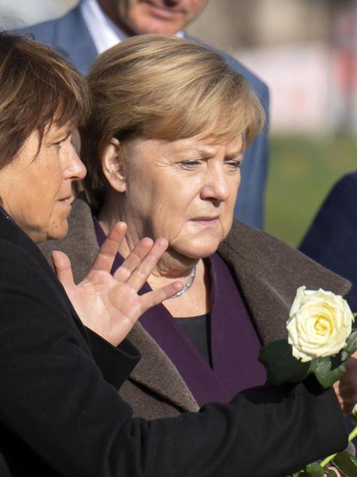 Die drei stehen nebeneinander, Merkel und Findeiß halten weiße Rosen in den Händen. Findeiß sagte etwas zu Merkel und gestikuliert mit der anderen Hand. Dahinter weitere Personen.
