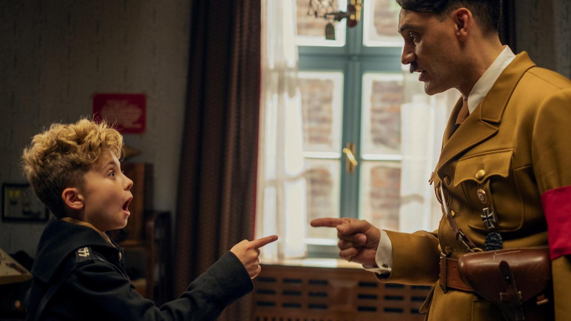 Szene aus dem Film "Jojo Rabbit": Ein Junge und ein Mann in der Verkleidung Hitlers zeigen mit Fingern aufeinander. Szene aus dem Film "Jojo Rabbit": Ein Junge und ein Mann in der Verkleidung Hitlers zeigen mit Fingern aufeinander.