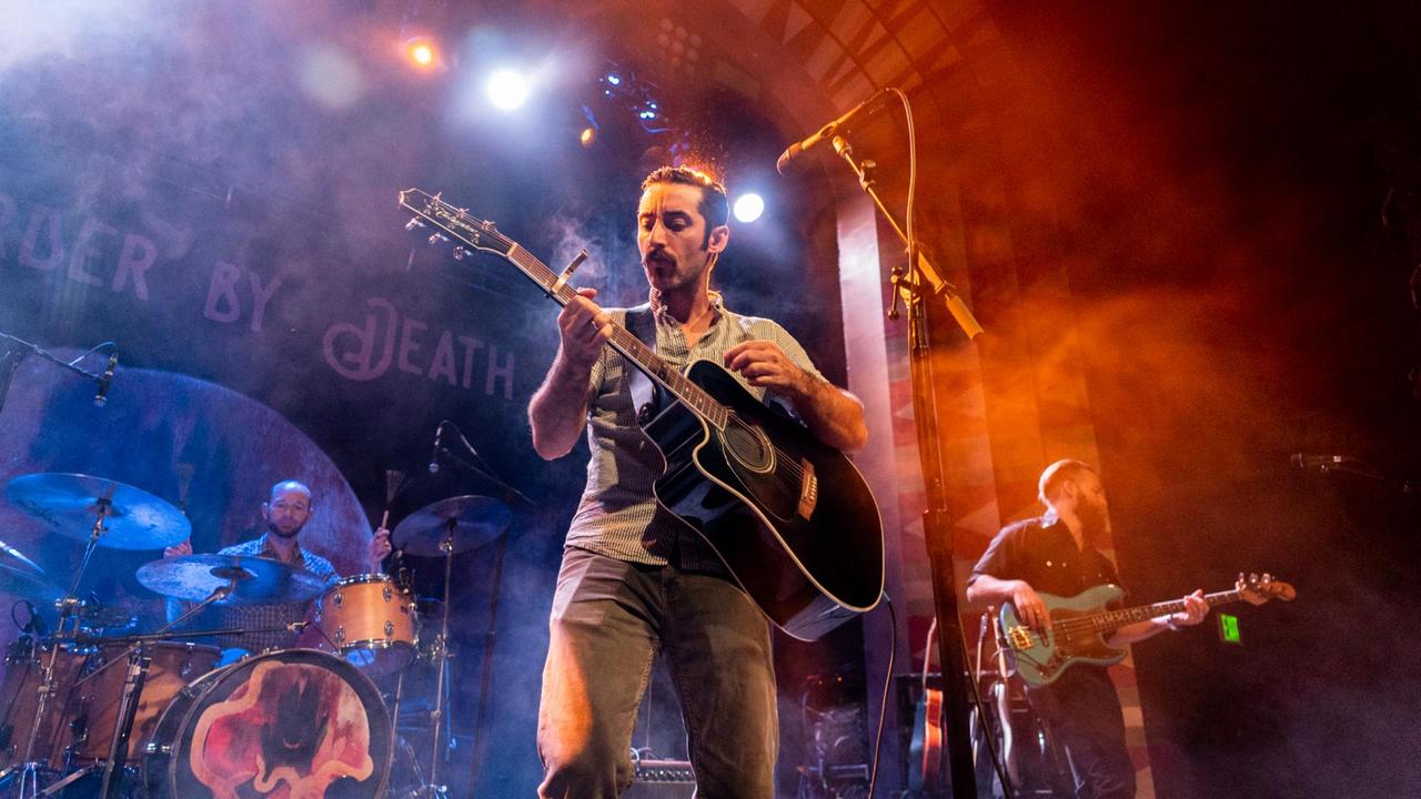 Zwei Männer mit Gitarre und ein Mann am Schlagzeug stehen auf einer bunt ausgeleuchteten Bühne. Zwei Männer mit Gitarre und ein Mann am Schlagzeug stehen auf einer bunt ausgeleuchteten Bühne.