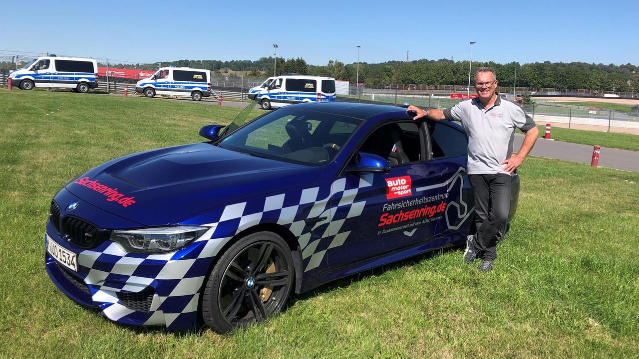 Auf einer grünen Wiese steht ein Mann im weißen T-Shirt vor seinem knallblauen Sportwagen. Im Hintergrund sieht man drei Polizeiwagen. Der Himel ist sommerlich strahlend blau. Auf einer grünen Wiese steht ein Mann im weißen T-Shirt vor seinem knallblauen Sportwagen. Im Hintergrund sieht man drei Polizeiwagen. Der Himel ist sommerlich strahlend blau.