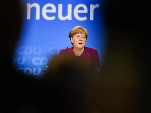Merkel steht auf der Bühne und spricht zu den Delegierten. Sie ist aus der Ferne fotografiert, der Blick auf sie wird durch die Schatten von Zuschauern eingeschränkt.