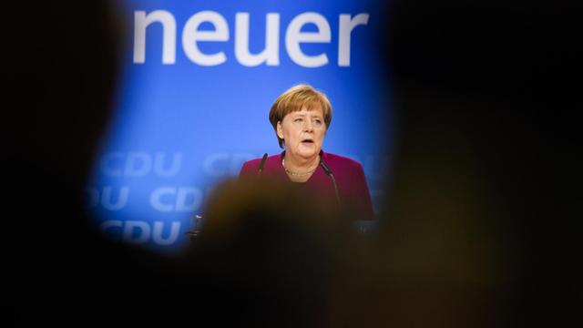 Merkel steht auf der Bühne und spricht zu den Delegierten. Sie ist aus der Ferne fotografiert, der Blick auf sie wird durch die Schatten von Zuschauern eingeschränkt.