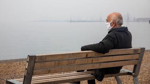 Mann mit Schutzmaske sitzt auf einer Bank an der Uferpromenade.