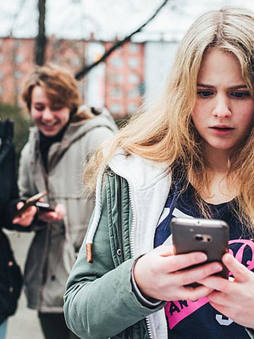 Ein Mädchen steht im Hof einer Schule und schaut auf ihr Smartphone, während im Hintergrund zwei andere Mädchen lachen.
