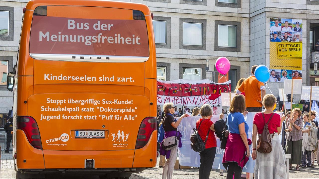 Ein orangener Bus zeigt die Aufschrift: "Stoppt übergriffigen Sex-Unterricht." Daneben stehen einige Demonstranten.