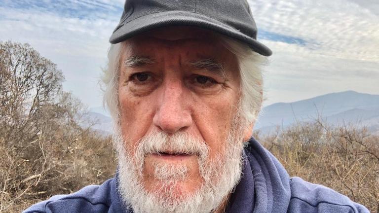 Ernesto Gonzalo Cáceres hat weißes Haar und trägt Vollbart. Er hat ein Basecap auf dem Kopf. im Hintergrund sind Bäume ohne Laub und eine Gebirgslandschaft zu sehen. Ernesto Gonzalo Cáceres hat weißes Haar und trägt Vollbart. Er hat ein Basecap auf dem Kopf. im Hintergrund sind Bäume ohne Laub und eine Gebirgslandschaft zu sehen.