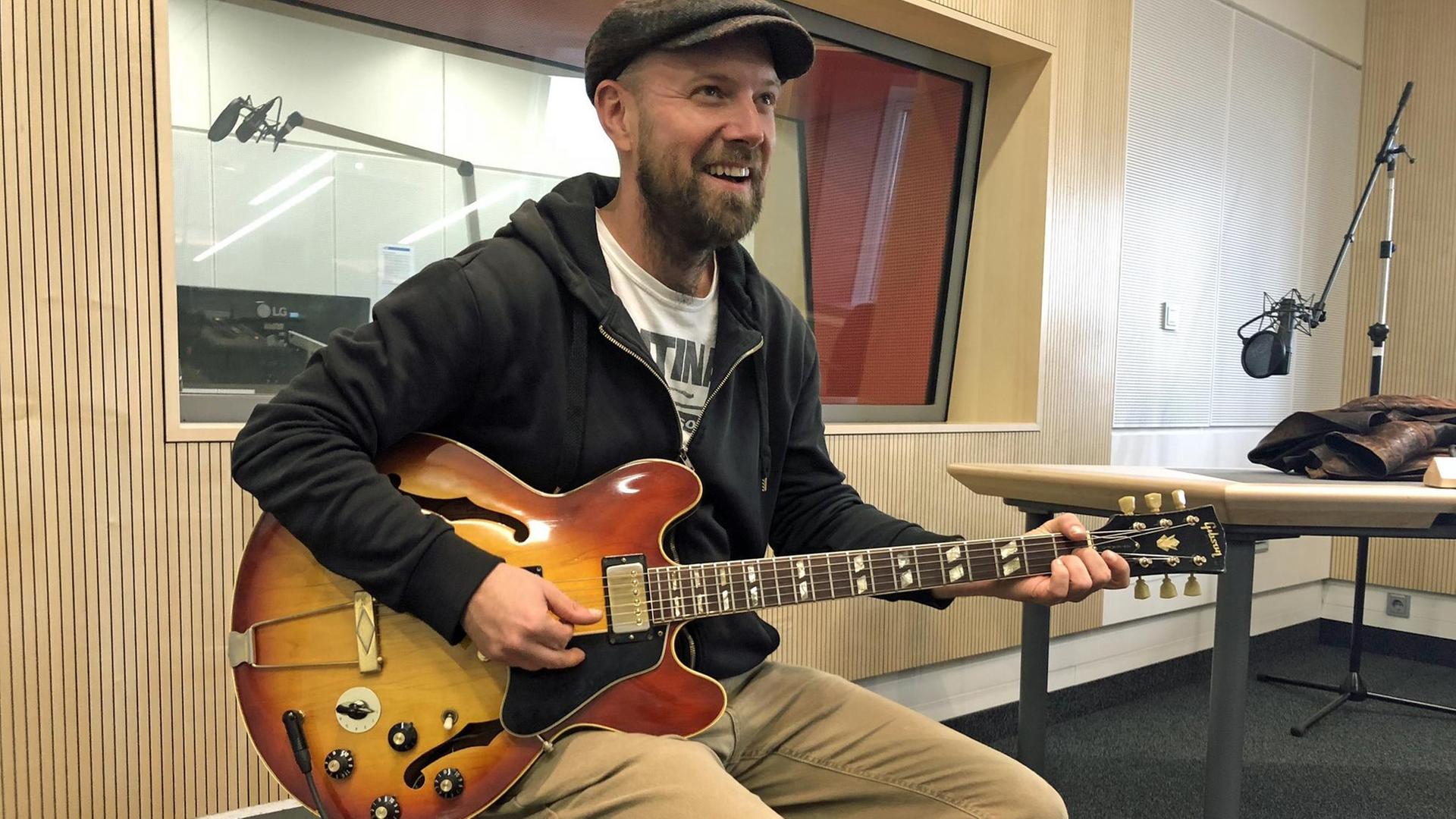 Ein Mann mit Kappe sitzt auf einer Kiste und hält eine halbakustische Gitarre in der Hand. Ein Mann mit Kappe sitzt auf einer Kiste und hält eine halbakustische Gitarre in der Hand.