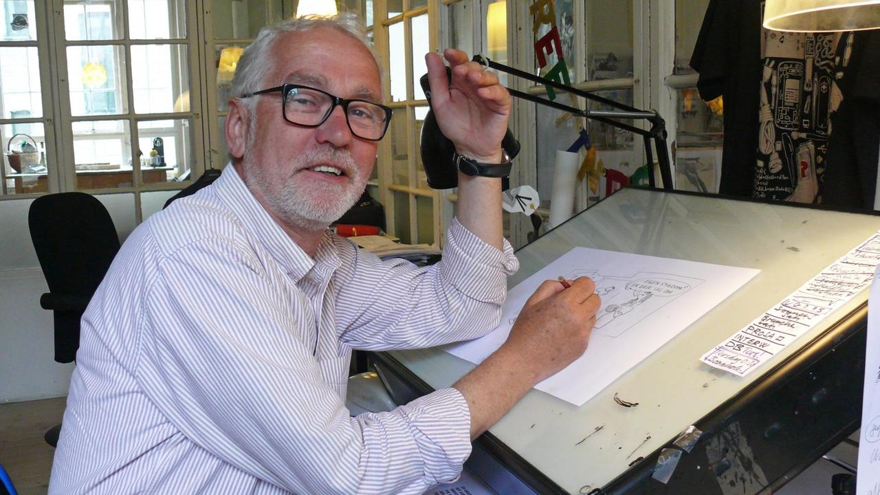 Der Karikaturist Lars Refn sitzt am Zeichentisch in seinem Atelier und schaut schelmisch lächelnd in die Kamera
