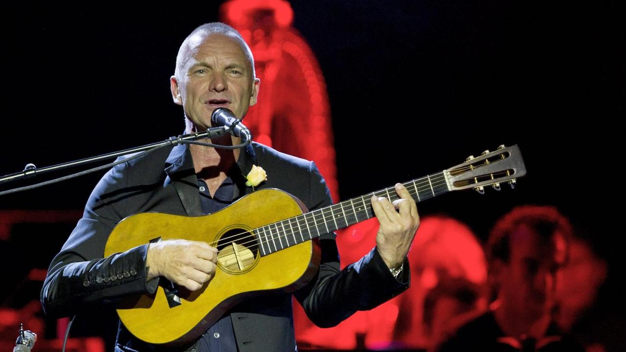 Ein Mann mit kurzen Haaren steht auf einer rot erleuchteten Bühne. Er spielt eine leuchtend gelbe Gitarre und singt dabei im Stehen vor einem Mikrophon.