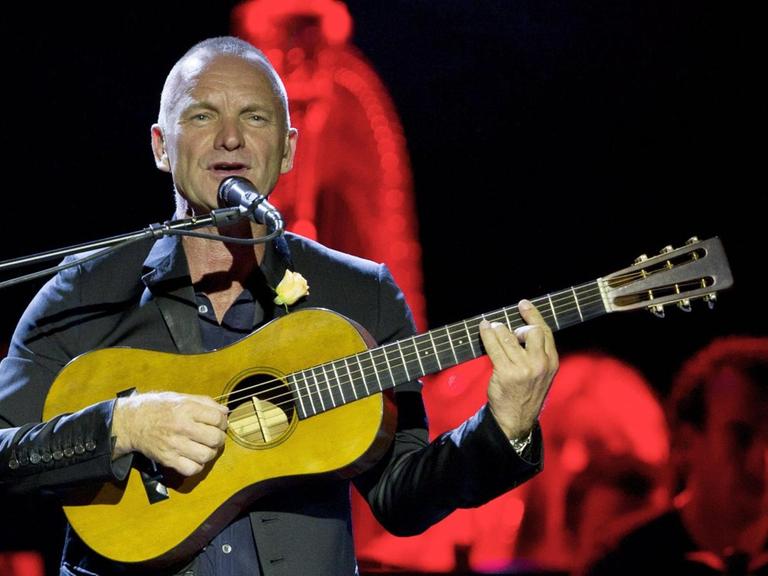 Ein Mann mit kurzen Haaren steht auf einer rot erleuchteten Bühne. Er spielt eine leuchtend gelbe Gitarre und singt dabei im Stehen vor einem Mikrophon.