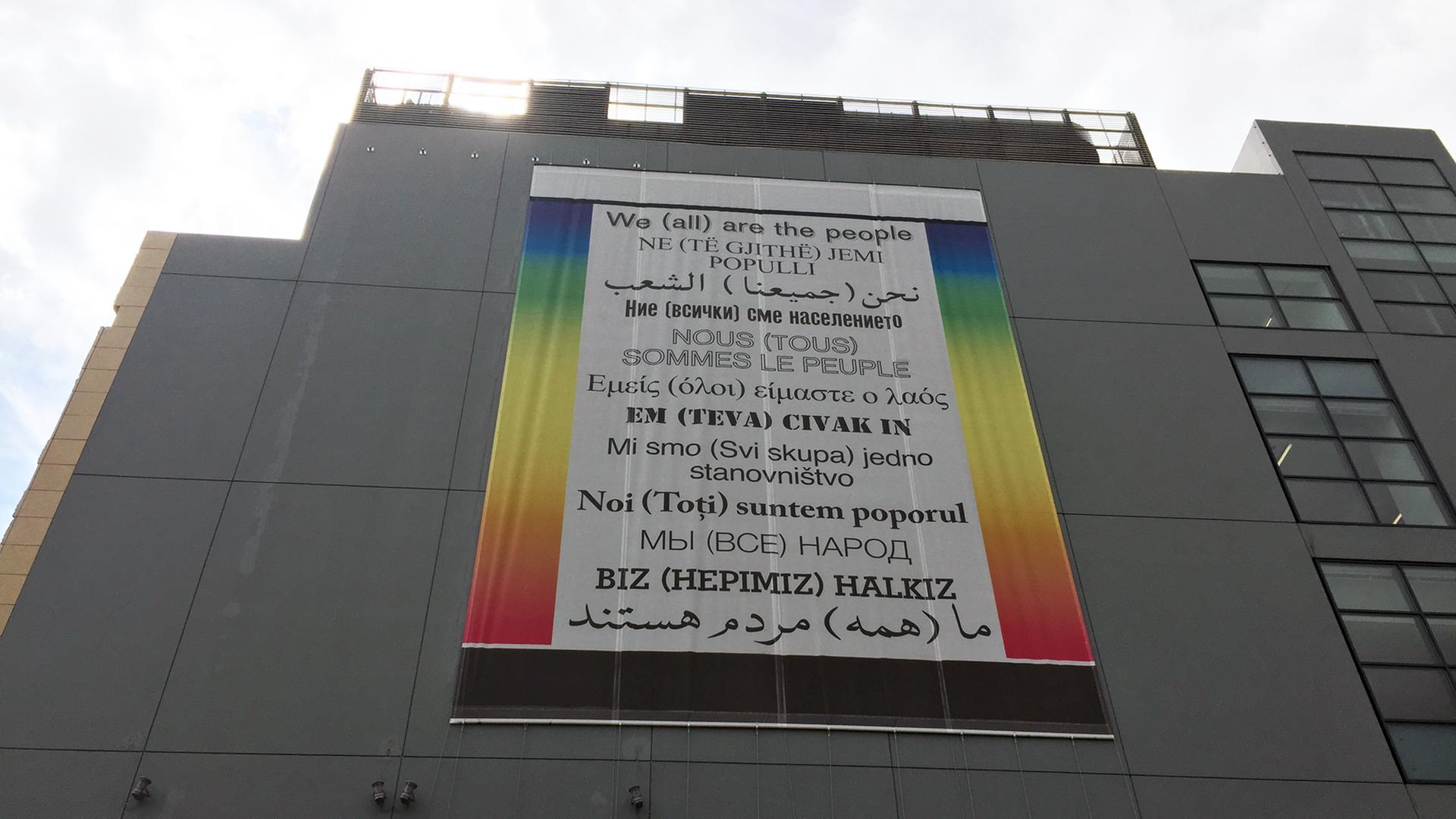 Plakat auf der Fassade des Museums für zeitgenössische Kunst (EMST) in Athen mit dem Satz "We (all) are the people" in unterschiedlichen Sprachen Plakat auf der Fassade des Museums für zeitgenössische Kunst (EMST) in Athen mit dem Satz "We (all) are the people" in unterschiedlichen Sprachen