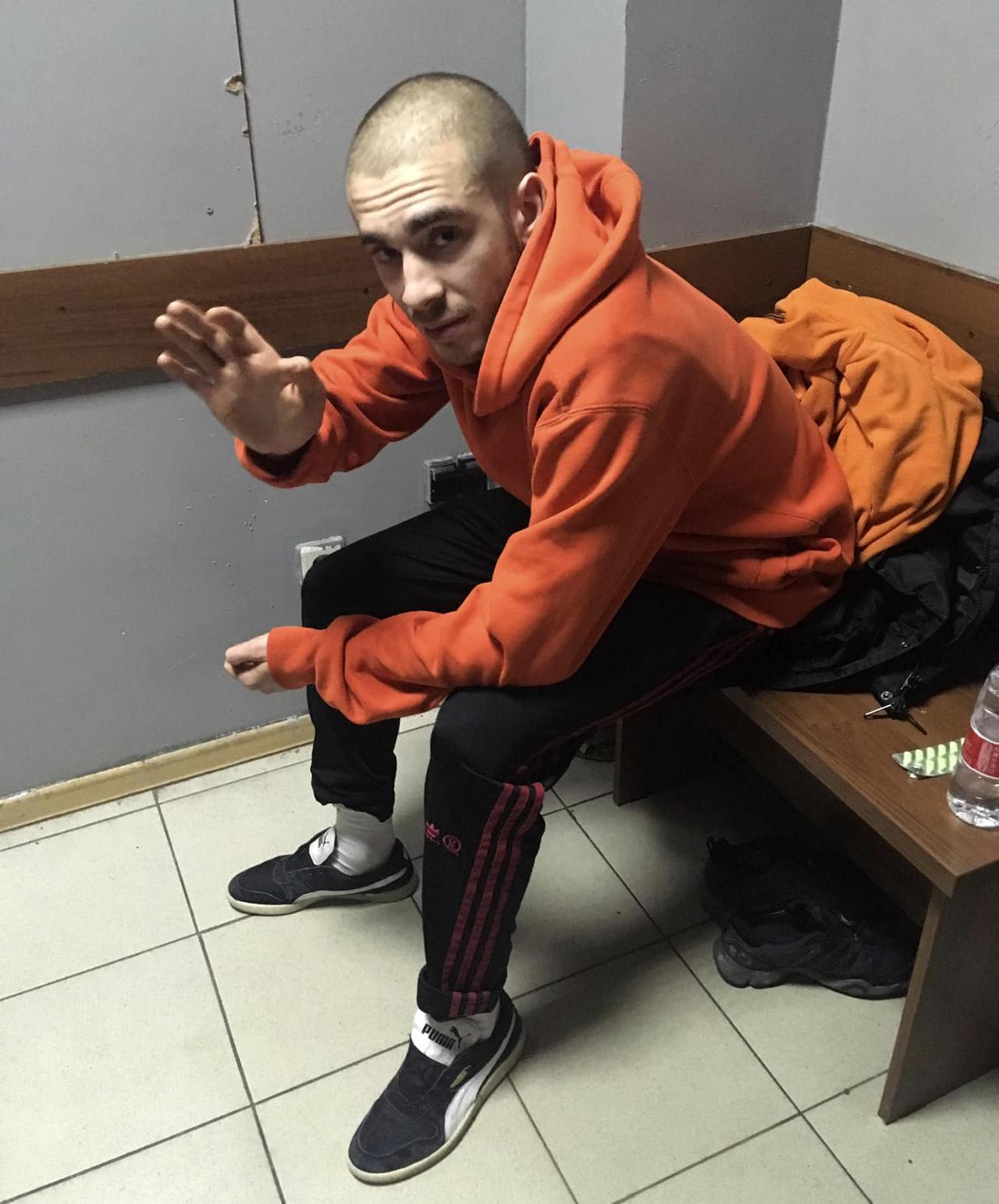 Der russische Rapper Husky sitzt in einem Gerichtsgebäude auf einer Bank und winkt mit der rechten Hand.