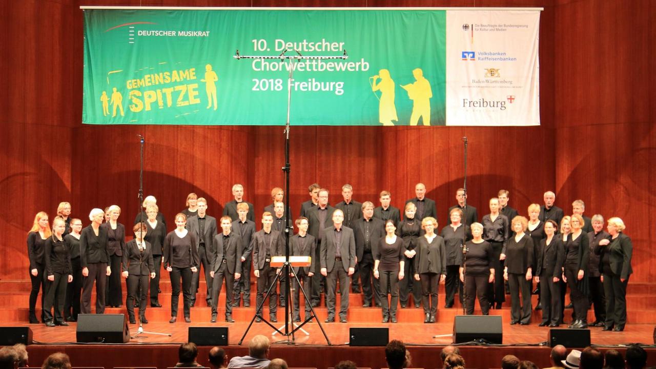 Der Chor steht beim auf der Bühne. Darüber ein großes Banner vom Chorwettbewerb in Freiburg. Der Chor steht beim auf der Bühne. Darüber ein großes Banner vom Chorwettbewerb in Freiburg.