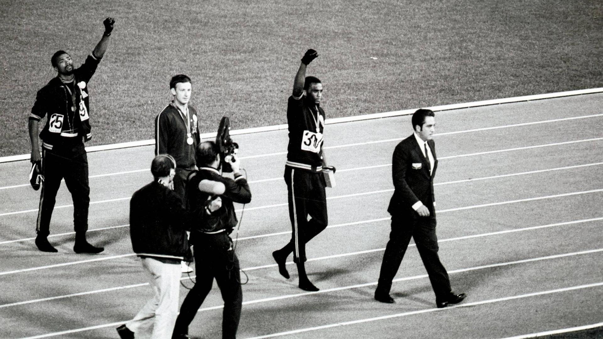 Ein Kamaramann fotografiert Tommie Smith und John Carlos, die kurz vorher die Black-Power-Faust bei der Siegerehrung in die Luft gereckt haben. Zwischen ihnen geht der Österreicher Peter Norman.