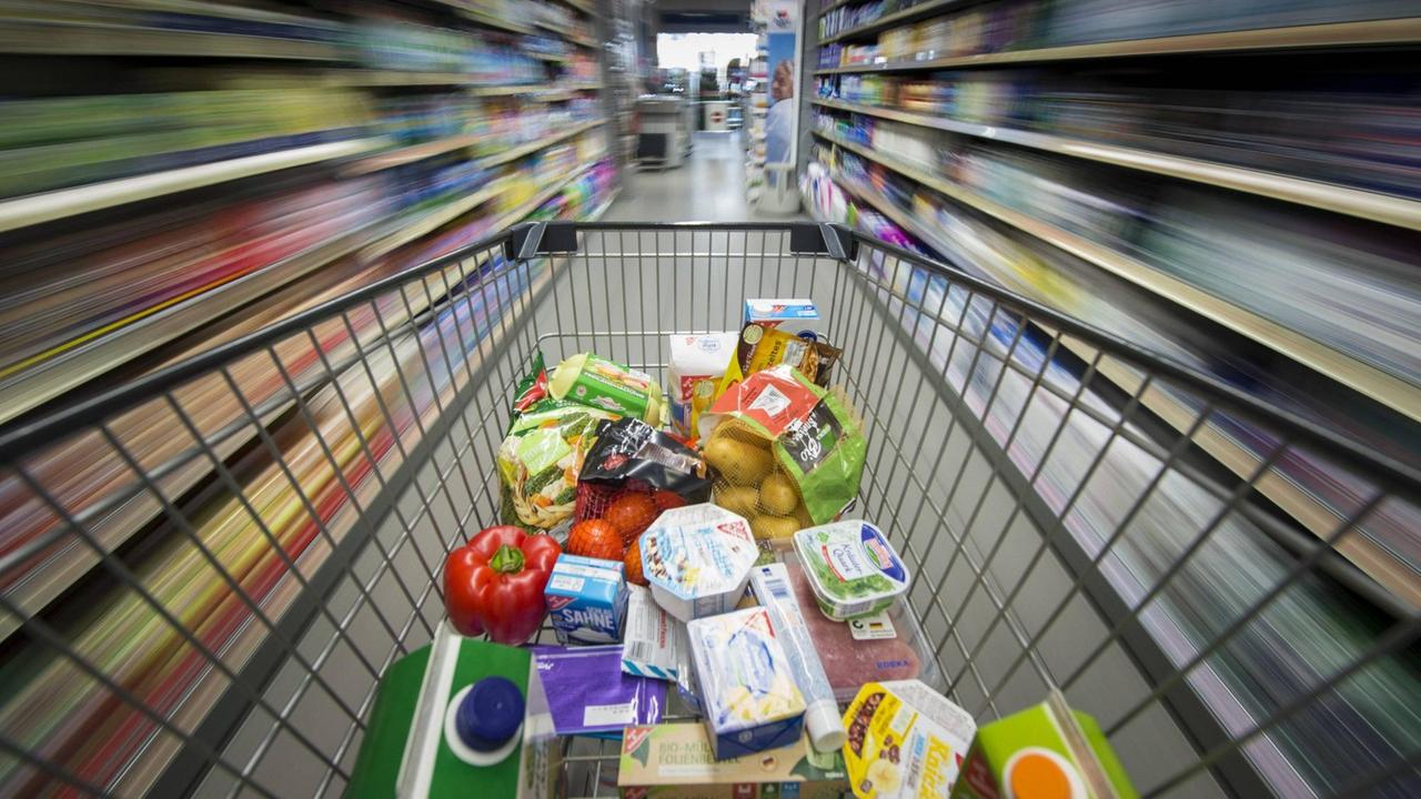 Ein Einkaufswagen mit verschiedenen Lebensmitteln wird durch die Gänge eines Supermarkts geschoben.