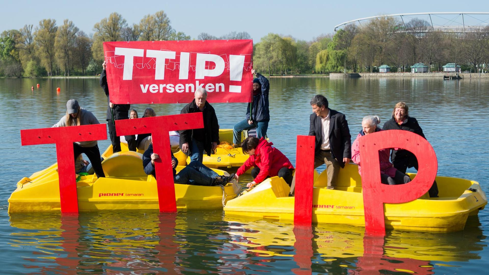 Die Aktivisten haben vier gelbe Tretboote zusammengefahren und halten übergroße rote TTIP-Buchstaben ins Wasser. Zwei halten ein rotes Transparent mit der Aufschrift "TTIP versenken!" hoch. Die Aktivisten haben vier gelbe Tretboote zusammengefahren und halten übergroße rote TTIP-Buchstaben ins Wasser. Zwei halten ein rotes Transparent mit der Aufschrift "TTIP versenken!" hoch.