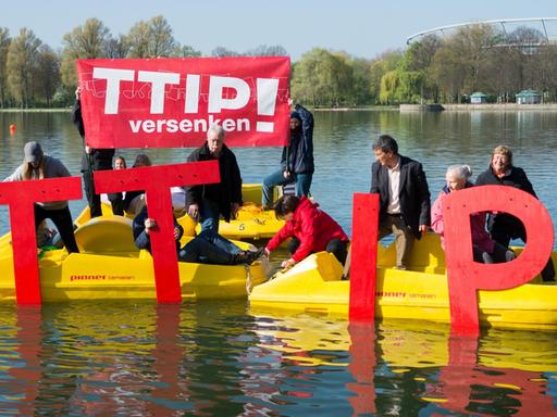 Die Aktivisten haben vier gelbe Tretboote zusammengefahren und halten übergroße rote TTIP-Buchstaben ins Wasser. Zwei halten ein rotes Transparent mit der Aufschrift "TTIP versenken!" hoch.