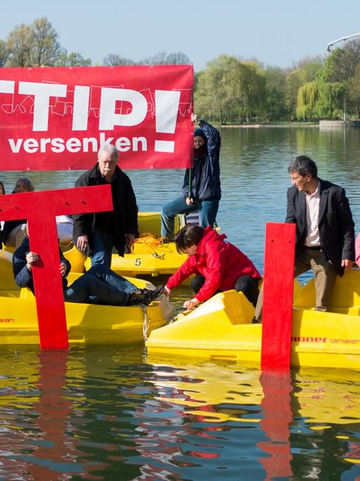 Die Aktivisten haben vier gelbe Tretboote zusammengefahren und halten übergroße rote TTIP-Buchstaben ins Wasser. Zwei halten ein rotes Transparent mit der Aufschrift "TTIP versenken!" hoch.