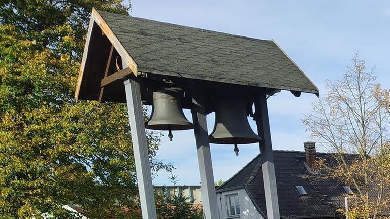 Zwei Glocken hängen in einem regengeschützten Holzgestell. Im Hintergrund sind Häuser der Gemeinde Jerche zu sehen. Zwei Glocken hängen in einem regengeschützten Holzgestell. Im Hintergrund sind Häuser der Gemeinde Jerche zu sehen.