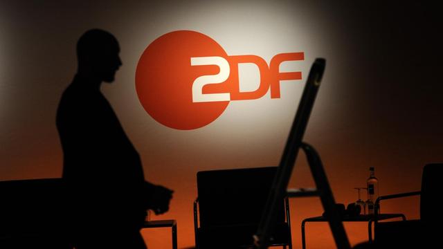 Ein Techniker steht neben einer Leiter vor einem ZDF-Logo. Auf einem Tisch sieht man Mineralwasser und Gläser.