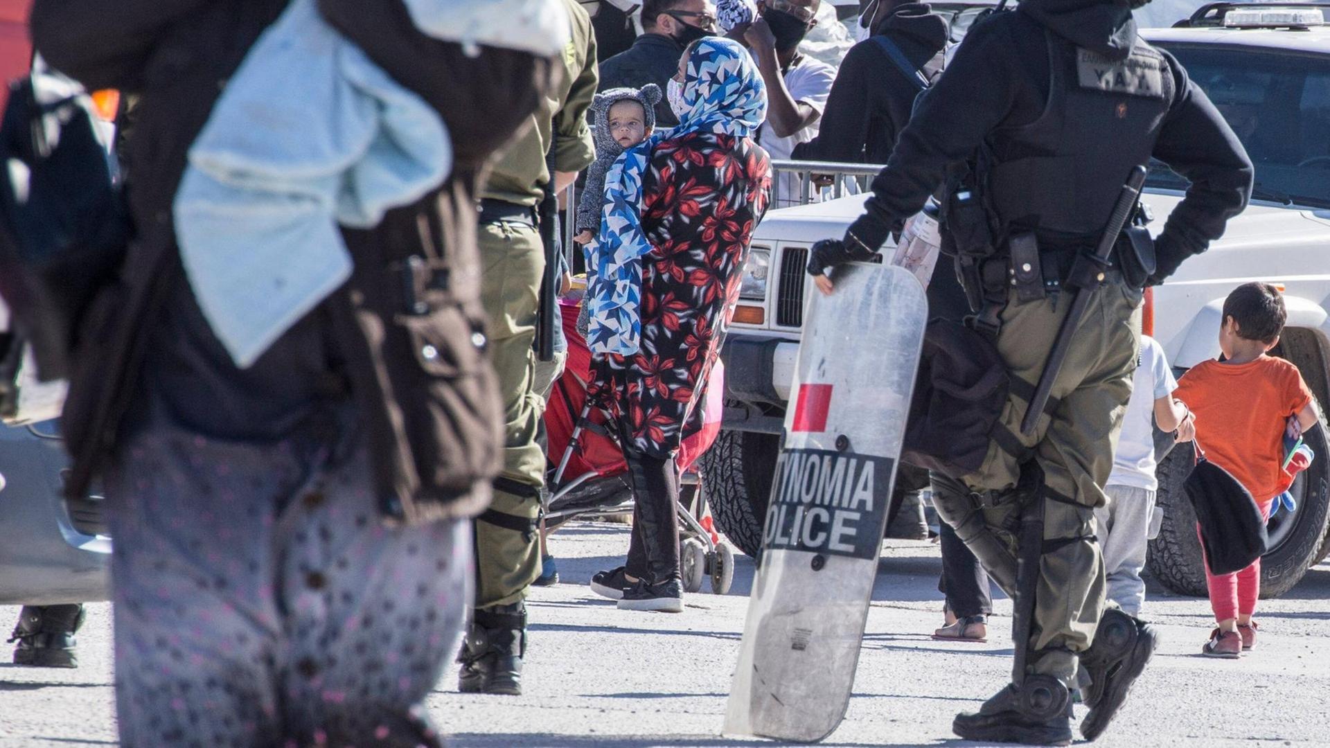Eine Asylsuchende mit ihrem Kind im November 2020 in der Warteschlange auf der griechischen Insel Lesbos vor dem Transport in das Flüchtlingslager Kara Tepe, bewacht von der griechischen Polizei Eine Asylsuchende mit ihrem Kind im November 2020 in der Warteschlange auf der griechischen Insel Lesbos vor dem Transport in das Flüchtlingslager Kara Tepe, bewacht von der griechischen Polizei