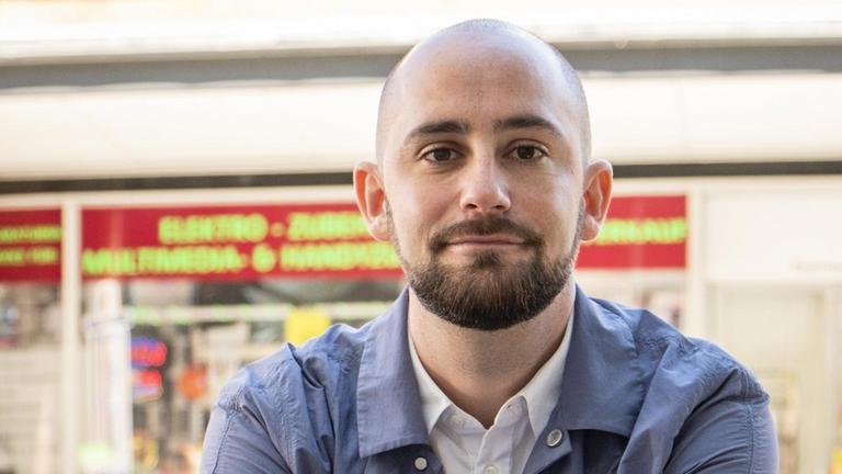 Philipp Staab, im weißen offenen Hemd und hellblauer Jacke, blickt freundlich in die Kamera. Im Hintergrund: die Frontscheibe eines Straßencafés, vor der Tische und Stühle in der Sonne stehen. Philipp Staab, im weißen offenen Hemd und hellblauer Jacke, blickt freundlich in die Kamera. Im Hintergrund: die Frontscheibe eines Straßencafés, vor der Tische und Stühle in der Sonne stehen.