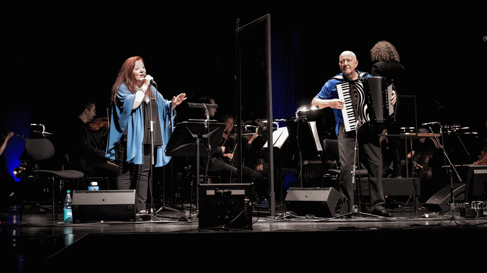 Die Sängerin Dima Orsho steht neben dem Akkordeonspieler Manfred Leuchter auf einer Bühne, auf der im Hintergrund ein Orchester sitzt.