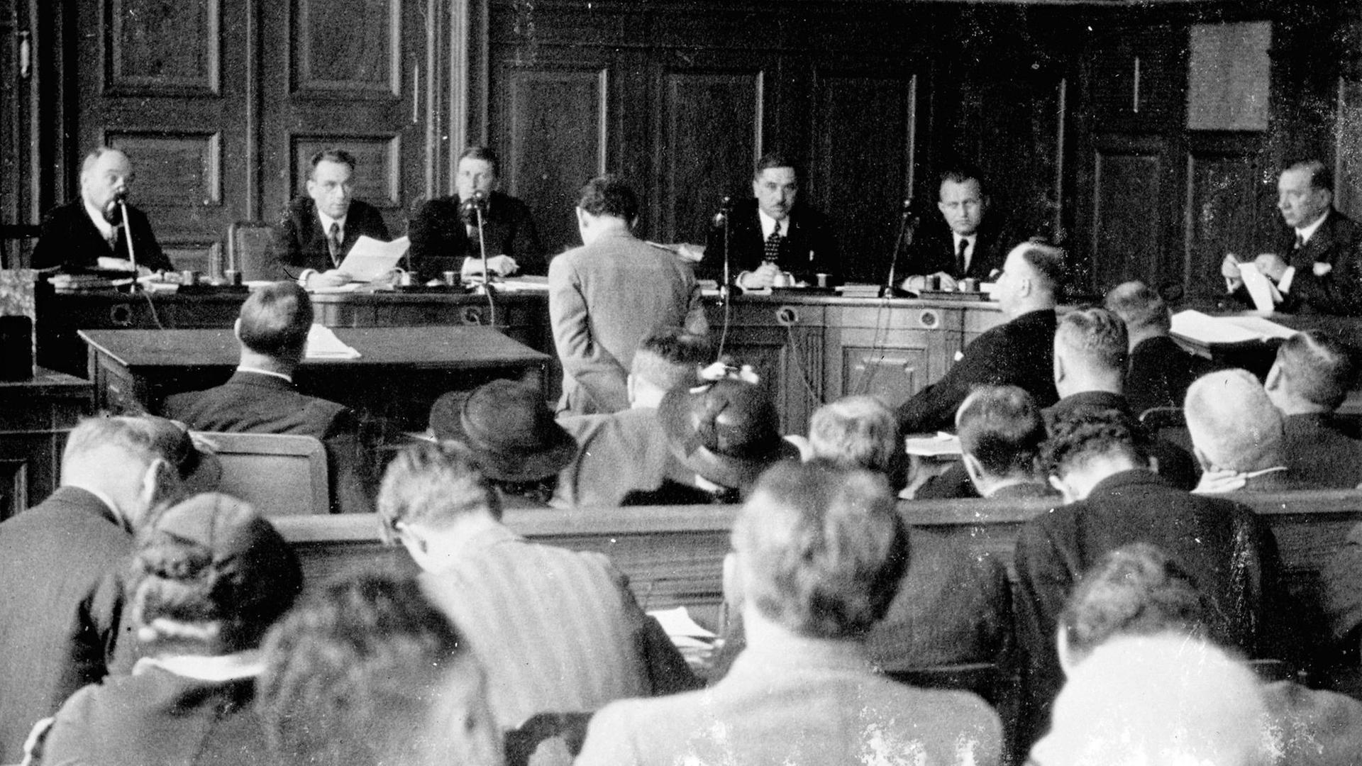Berthold Jacob (heller Anzug, stehend) und sein Kidnapper Hans Wesemann (dunkler Anzug, rechts sitzend) in einem Gerichtssaal in Basel im Mai 1936