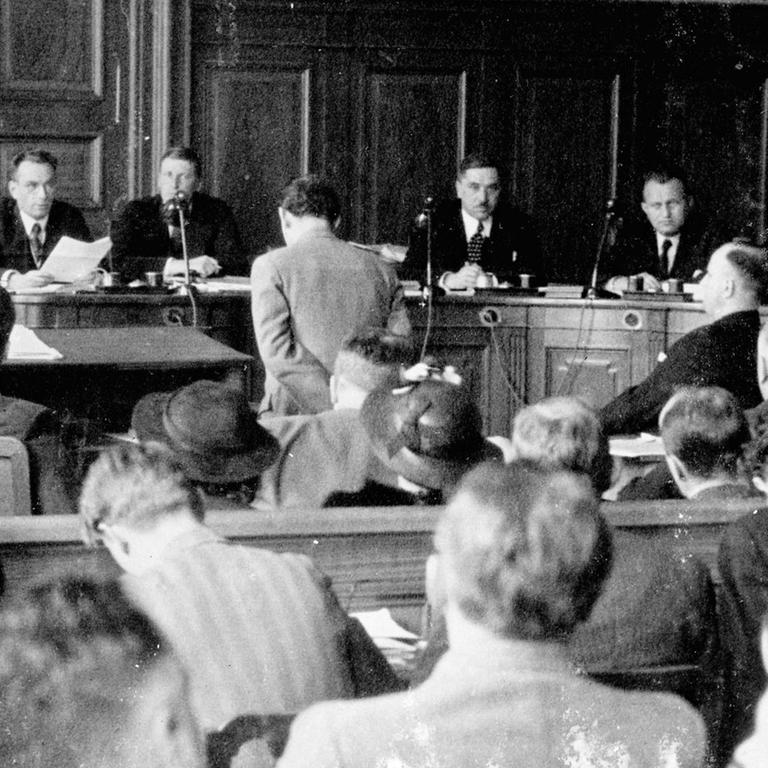 Berthold Jacob (heller Anzug, stehend) und sein Kidnapper Hans Wesemann (dunkler Anzug, rechts sitzend) in einem Gerichtssaal in Basel im Mai 1936