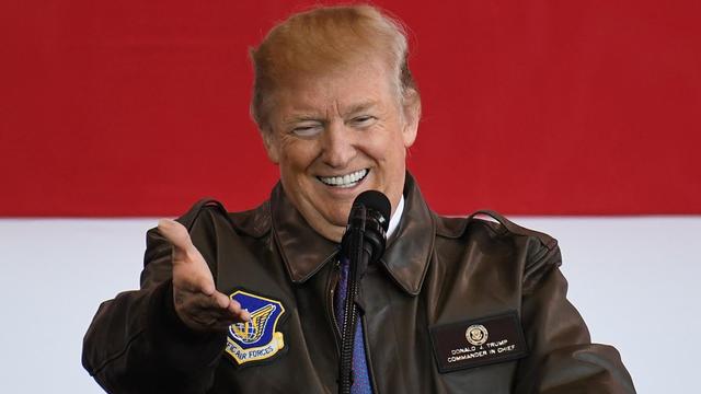 Trump trägt eine Pilotenjacke mit dem Aufnäher "Commander in Chief". Er steht vor einer riesigen US-Flagge, lacht und zeigt ins Publikum.
