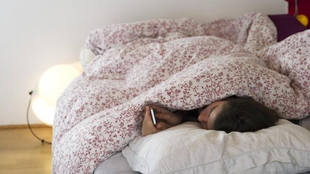 Eine Frau liegt im Bett und blickt auf ihr Mobiltelefon. Die Kommunikation über eine App soll in Zukunft bei Depressionen Lösungsansätze bieten.