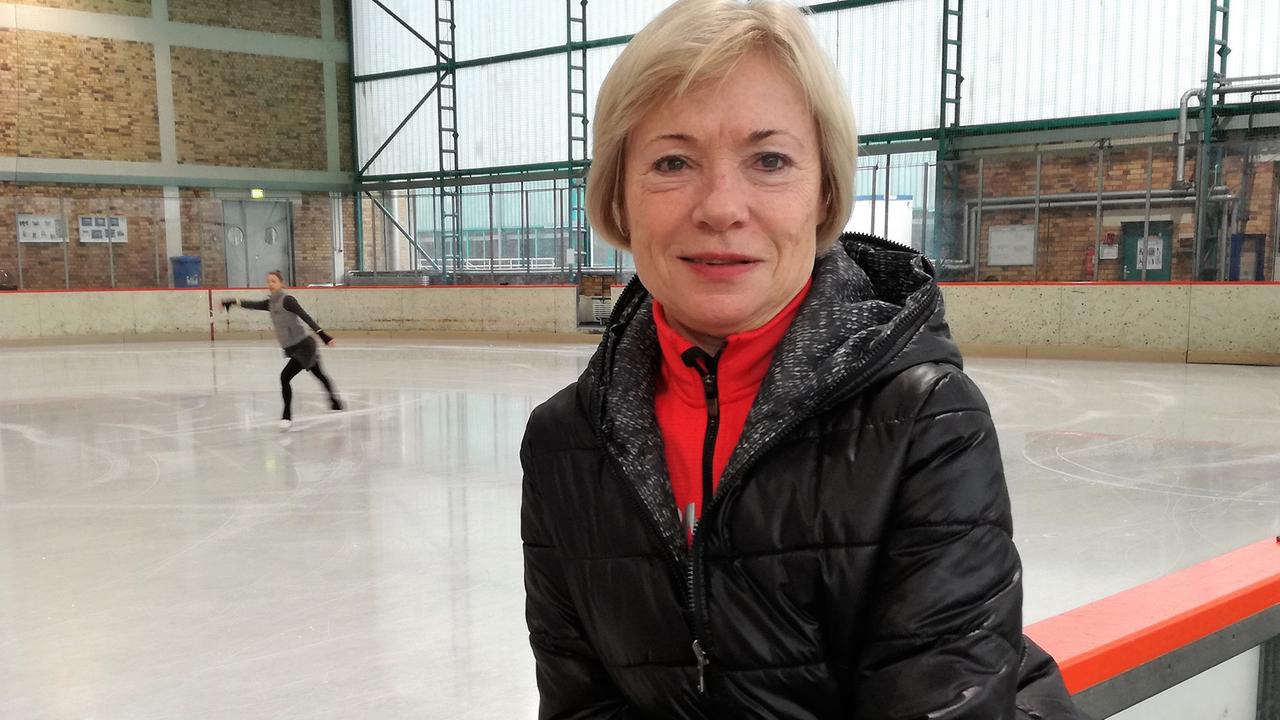 Eiskunstlauftrainerin Romy Österreich steht in einer Eiskunstlaufhalle, im Hintergrund läuft ein Mädchen Schlittschuh.