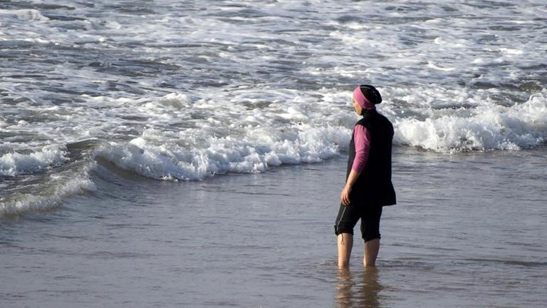 Eine Frau in Burkini steht mit den Füßen im Wasser und schaut aufs Meer.