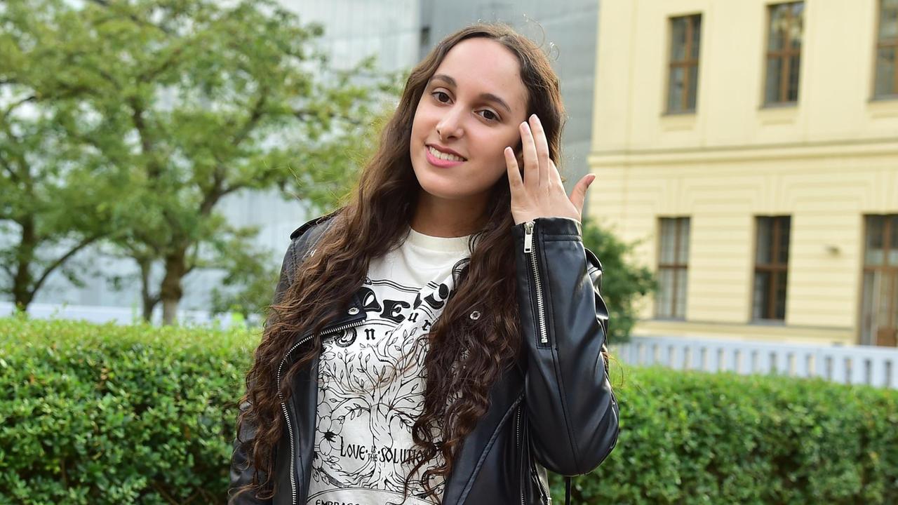 Die Rapperin und Songwriterin Sharon steht mit schwarzer Lederjacke im Garten des Jüdischen Museums Berlin und streicht sich die langen braunen Locken aus dem Gesicht.