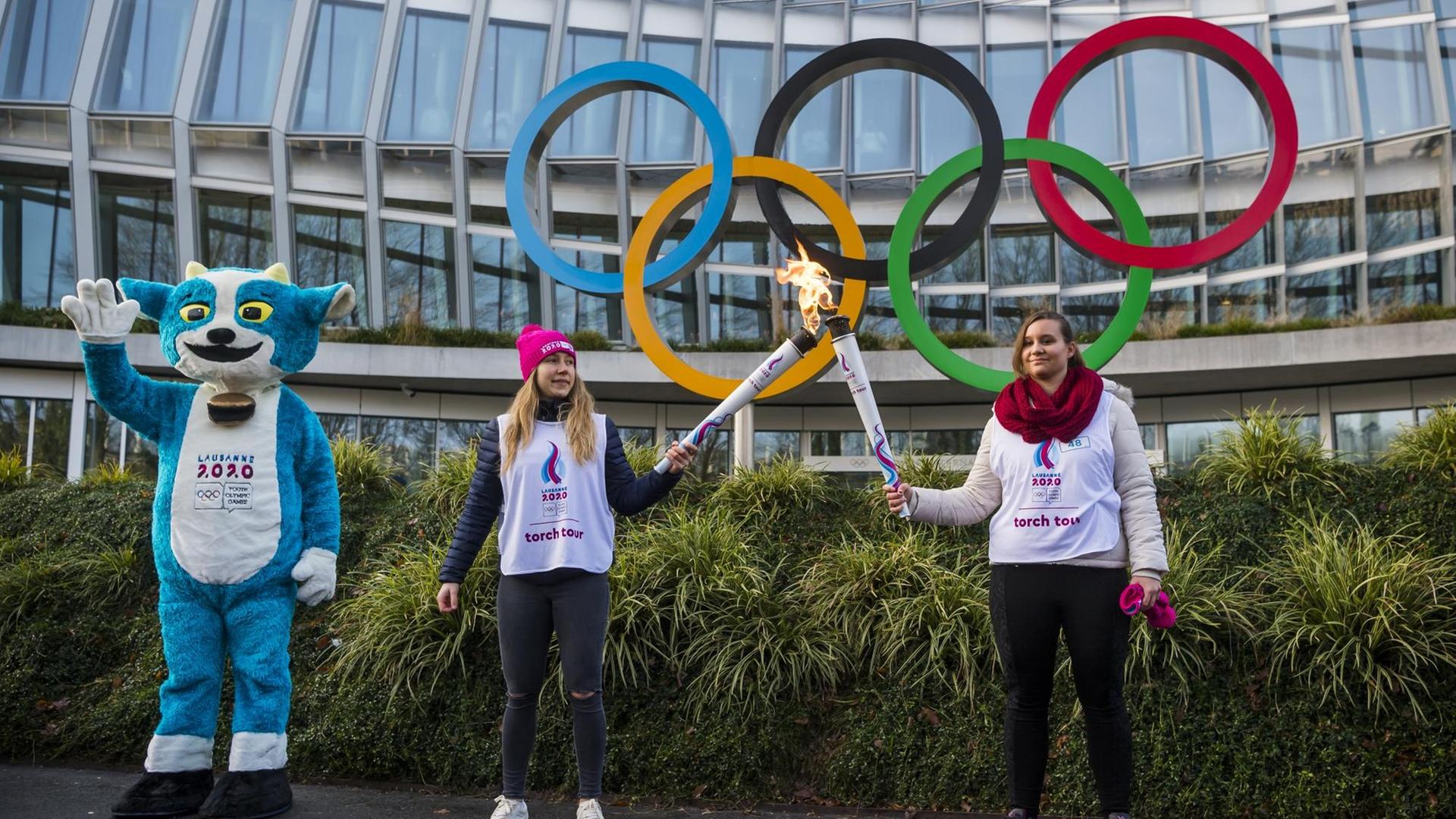 Schweiz, Lausanne: Yodli (l-r), das Maskottchen der Olympische Jugend-Winterspiele 2020, steht mit Christelle und Aurore, die die olympischen Flamme während einer Fackeltour halten, vor den Olympischen Ringen am Olympischen Haus. Schweiz, Lausanne: Yodli (l-r), das Maskottchen der Olympische Jugend-Winterspiele 2020, steht mit Christelle und Aurore, die die olympischen Flamme während einer Fackeltour halten, vor den Olympischen Ringen am Olympischen Haus.