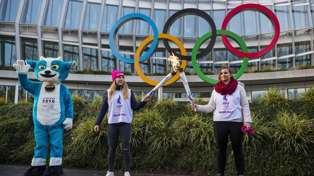 Schweiz, Lausanne: Yodli (l-r), das Maskottchen der Olympische Jugend-Winterspiele 2020, steht mit Christelle und Aurore, die die olympischen Flamme während einer Fackeltour halten, vor den Olympischen Ringen am Olympischen Haus.