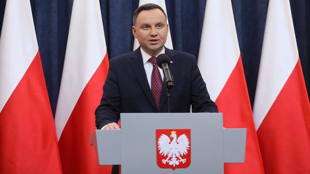 Duda steht an einem Rednerpult mit dem polnischen Adler und spricht. Hinter ihm vier polnische Flaggen vor einem blauen Vorhang.